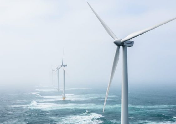 Modern wind turbines on the North Sea coastline under a bright overcast sky, with waves and distant turbines in the background.